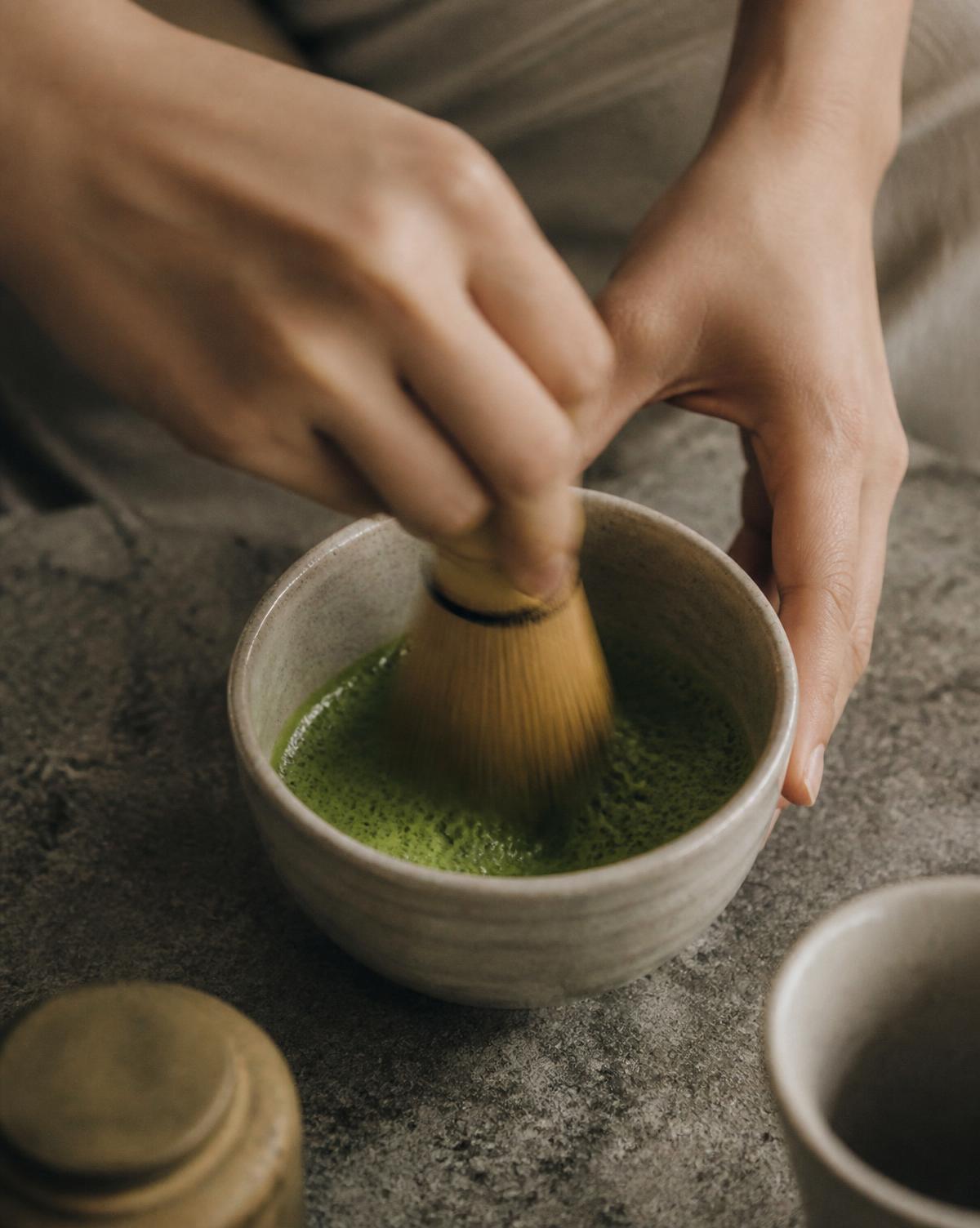 Two hands whisking matcha with a bamboo chasen in a ceramic bowl